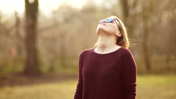 Lovely young blonde in glasses puts her face to the spring sun. The girl laughs cheerfully. She arranges his hands and goes towards the wind. Unity with nature. Enjoy the spring heat — Stock Video