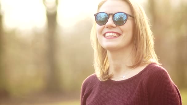 Uma mulher de meia-idade bonito olha para a câmera estritamente e benevolentemente sorrindo. Ela usa óculos escuros. Uma menina descansa no parque da cidade no início da primavera. Pôr do sol — Vídeo de Stock