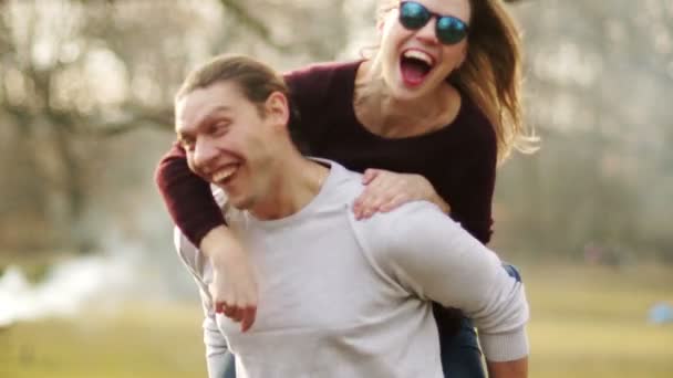 Schöner Kerl, der seiner Freundin eine Huckepackfahrt gibt, beide haben Spaß am Ende mit Kirschbäumen im Hintergrund — Stockvideo