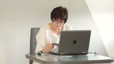 Yaşlı bir kadın ile bir dizüstü bilgisayar ve bir banka kartı faturaları ödüyor. Online ödeme