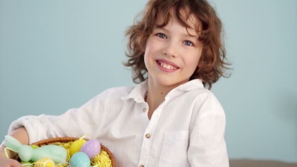 Σγουρά κοκκινομάλλα παιδί σε ένα λευκό πουκάμισο με μια φιγούρα από ένα λαγουδάκι του Πάσχα και ένα σύνολο Πασχαλινά αυγά σε ένα καλάθι. Ένα αγόρι γελάει χαρωπά. Αργή κίνηση. — Αρχείο Βίντεο