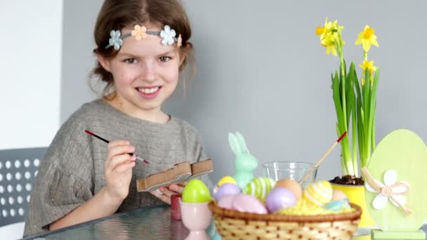 Brown-eyed girl z wieniec na głowie przygotowuje się do Świąt Wielkanocnych. Dziecko patrzy w kamerę i śmieje się wesoło. W jej rękach farby i pędzla. Na stole jest wielkanocnym koszyku i bukiet — Wideo stockowe