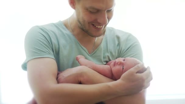 Молодой отец атлета строит обнаженного новорожденного ребенка на своих руках. Мужчина гладит его по голове, ребенок машет ручкой и поворачивает голову, оглядывается. День отца — стоковое видео