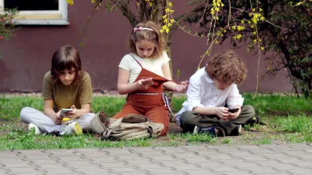 Deux frères et une sœur utilisent des smartphones assis sur l'herbe. Socialiser dans les réseaux sociaux. Dépendance réseau. Organisation des loisirs des écoliers — Video