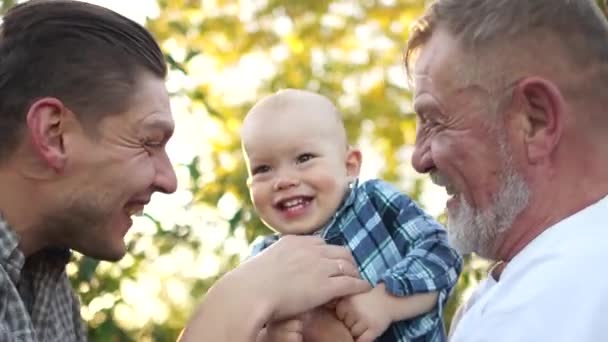 Niño pequeño, su padre y su abuelo se divierten en el jardín de verano. Padre hace cosquillas a su hijo, el bebé se sienta en los brazos de su abuelo y se ríe alegremente — Vídeo de stock