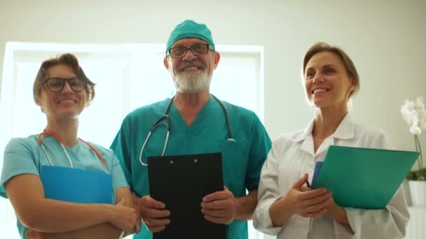 Dokter en twee verpleegsters poseren voor de camera. Artsen lachen terwijl ze voor het raam staan en medische documenten in hun handen houden. Internationale verpleegdag — Stockvideo