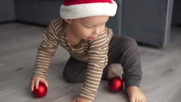 Peuter in een Santas fleece hoed speelt met een rode kerstbal. Vrolijk Kerstfeest en Gelukkig Nieuwjaar — Stockvideo