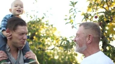 Mutlu bebek babasının omuzlarında oturuyor, büyükbaba yakınlarda duruyor, bebek eğiliyor ve herkes neşeyle gülüyor. Açık havada portre