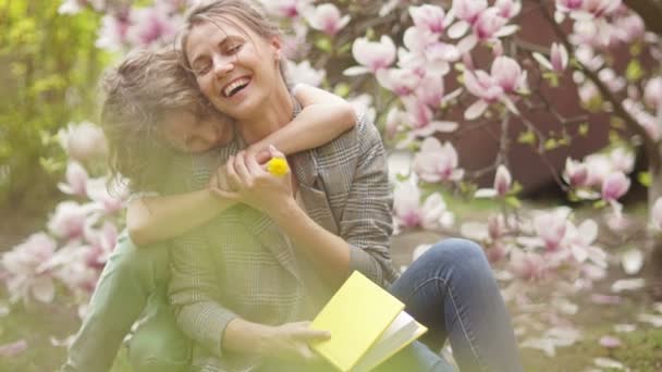 꽃 이 핀 마뇰 리아 나무 옆에 엄마와 함께 있는 사랑 스러운 어린 소년. 어머니 날, 봄 방학 — 비디오