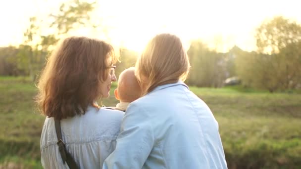 Couple de lesbiennes avec un bébé dans les bras pour une promenade dans le parc d'automne. Deux femmes se tiennent debout embrassant et regardant le coucher du soleil, vue arrière — Video