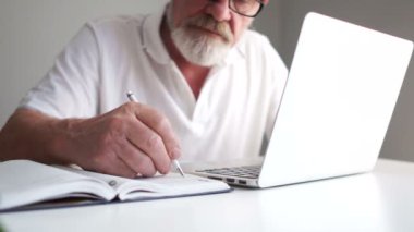 Gri sakallı bir iş adamı ofiste dizüstü bilgisayar kullanıyor. Not defterindeki kalemle not alıyor. Uzaktan istihdam, iş yerindeki yaşlı vatandaş, bilgisayardaki adam