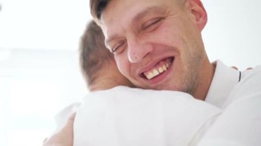 Yaşlı baba yetişkin oğluna sarılıyor. Neşeli bir buluşma, baba oğul, babalar günü, beyazlar içindeki iki adam kapalı alanda birbirlerine sarılıyorlar.