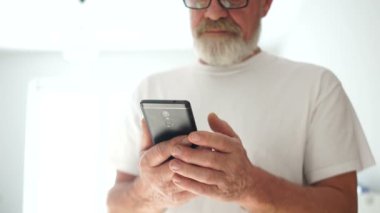 Gözlüklü ve beyaz tişörtlü gri sakallı bir adamın portresi akıllı telefon kullanıyor. Akıllı telefondan fotoğraf çeken gülümseyen olgun bir adam. Modern emekliler