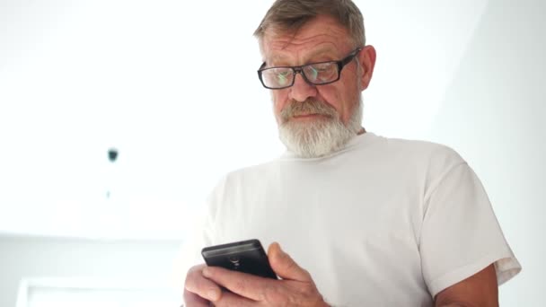 Un hombre de barba gris maduro con una camiseta blanca y gafas marca un número en un teléfono inteligente y comienza una conversación. Viejo y smartphone, tecnología moderna retirada — Vídeos de Stock