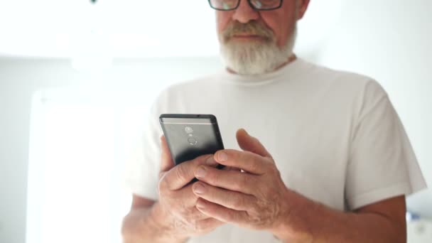 Un retrato cercano de un hombre de barba gris con gafas y una camiseta blanca está usando un teléfono inteligente. Sonriendo hombre maduro hojeando a través de fotos en el teléfono inteligente. Pensionista moderno — Vídeo de stock