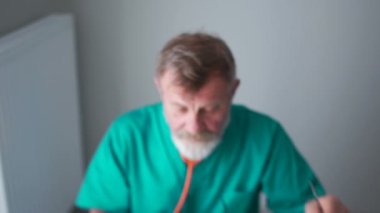 Yaşlı bir doktor, yeşil bir tıbbi önlük giyen profesör hasta kayıtlarını dolduruyor ve gülümseyen kameraya bakıyor.