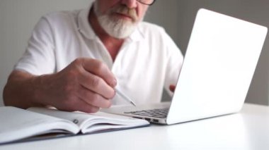 Beyaz tişörtlü ve gri sakallı olgun bir adam dizüstü bilgisayar kullanarak internette çalışırken kalemle not alıyor. Ofis çalışanı, bölüm başkanı, iş adamı konsepti