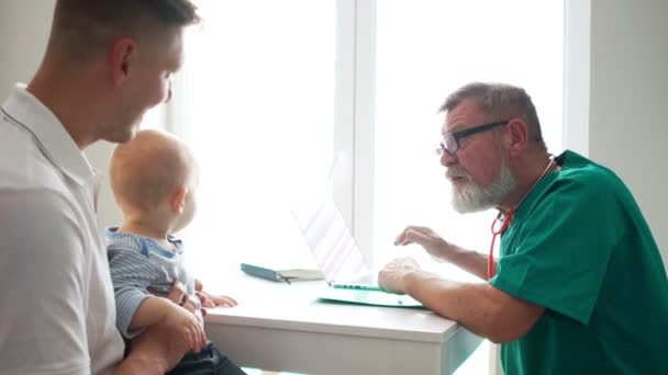 Маленький ребенок с отцом на приеме у педиатра. Пожилой педиатр осматривает ребенка в своем кабинете, слушает дыхательный стетоскоп — стоковое видео