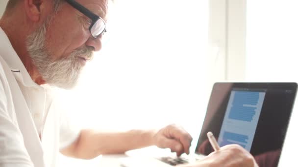 Senior man arbetar med bärbar dator hemma. Äldre farfar bär glasögon och arbetar på laptop i vardagsrummet. Äldre medborgares hobbyer och fritid — Stockvideo