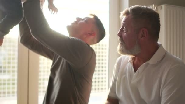 Abuelo con hijo y nieto sonriendo sentado en el sofá. El hijo adulto levanta al nieto por encima de la cabeza. tres generaciones de hombres de la misma familia feliz. Día de los Padres, Retrato interior — Vídeos de Stock