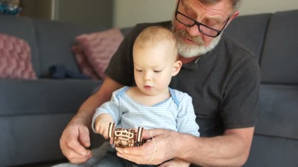 Dědeček a vnuk si hrají s dřevěnou hračkou, objímají se a usmívají a odpočívají spolu doma. Šťastný dědeček sedět na gauči těšit trávit čas s předškolním vnukem doma — Stock video