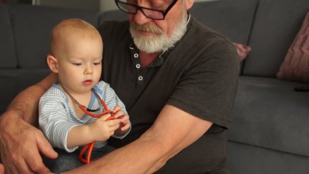 El abuelo y el nieto están jugando al médico con el fonendoscopio, abrazándose y sonriendo mientras descansan juntos en casa. Feliz abuelo sentarse en el sofá disfrutar de pasar tiempo con el nieto preescolar en casa — Vídeo de stock