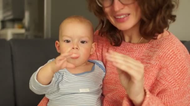 Lycklig mamma leker med sitt barn på soffan hemma. Spädbarn sitter på mammor och leker och tittar på henne. Ung leende kvinna barnvakt söt småbarn — Stockvideo