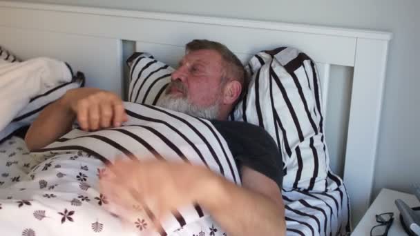 Een oudere man met een baard wordt wakker en komt 's morgens uit bed, strekt zich uit, houdt zijn zere schouder vast — Stockvideo