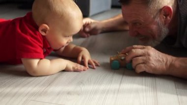 Dedem küçük torunuyla oyuncak tahta bir arabayla oynuyor, dedesiyle ilgileniyor ve tatlı bir torunu sıcak zeminde yatıyor, evde eğleniyor, ısıtma konseptini kapatıyor.