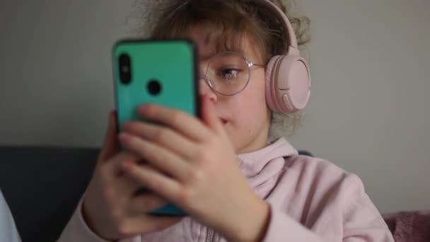 Portrait d'une jolie adolescente en lunettes et avec écouteurs sans fil. Dans les mains d'une fille tenant un smartphone. Un enfant écoute de la musique, un adolescent et de la musique — Video