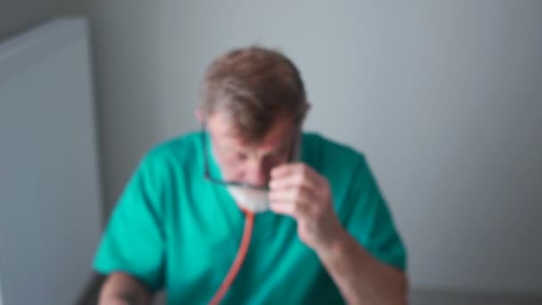 Nahaufnahme des Therapeuten. ein älterer Arzt, Professor im grünen Arztkittel füllt eine Patientenakte aus und blickt lächelnd in die Kamera — Stockvideo