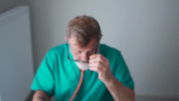 Close portrait of therapist. An elderly doctor, professor wearing a green medical gown fills out a patients medical record and looks at the camera smiling — Stockvideo