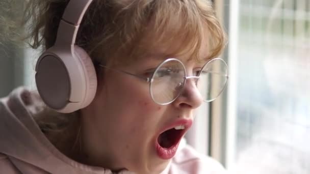 Chica adolescente con auriculares rosados y gafas aburridas mientras se sienta en casa cerca de la ventana. Chica bosteza cubriendo su boca con su mano — Vídeos de Stock