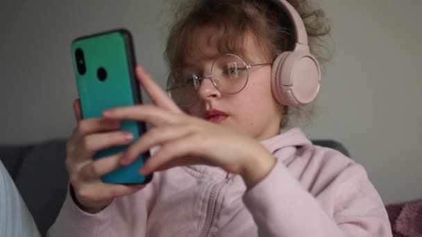 Skolflicka i glasögon med telefon lyssnar på musik via trådlösa hörlurar. Nära porträtt av lockigt söt tonåring flicka, ungdom subkultur — Stockvideo