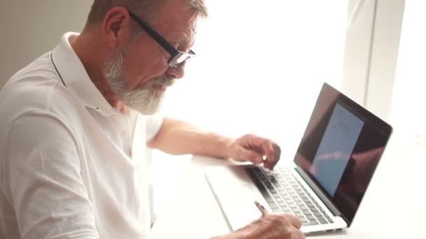 Der graubärtige Schriftsteller schreibt mit Hilfe eines Laptops ein Buch. Älterer Mann arbeitet zu Hause mit Laptop — Stockvideo