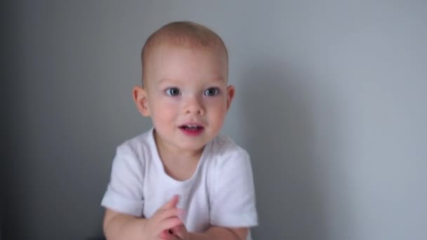 一个穿着白色T恤的两岁蓝眼睛男孩的画像。 可爱的小男孩看着摄像机，拍了拍他的手。 有趣的视频，快乐的童年 — 图库视频影像