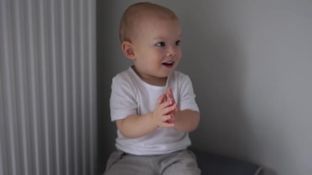 Lindo bebé de dos años con una camiseta blanca sentada en una silla y aplaudiendo en el interior. Feliz infancia. — Vídeos de Stock