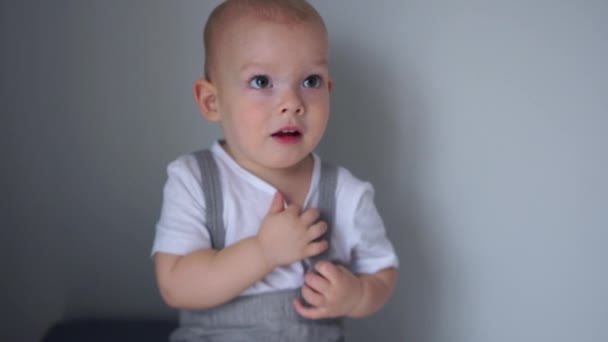 Retrato próximo de um menino de dois anos de idade de olhos azuis. A criança grita algo, crianças emoções. Criança vestida com uma camiseta branca e calças cinza — Vídeo de Stock