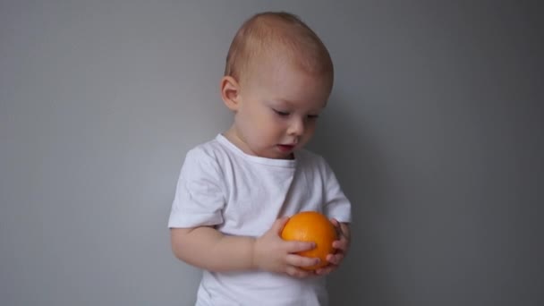 Hält das Kind eine Orange in den Händen, ein nahes Porträt eines zweijährigen blauäugigen Jungen. Gesunde Ernährung — Stockvideo