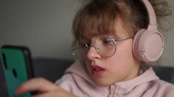 Κοντινό πορτρέτο του σγουρού χαριτωμένο έφηβος κορίτσι, υποκουλτούρα της νεολαίας. Μαθήτρια με γυαλιά και τηλέφωνο ακούει μουσική μέσω ασύρματων ακουστικών — Αρχείο Βίντεο