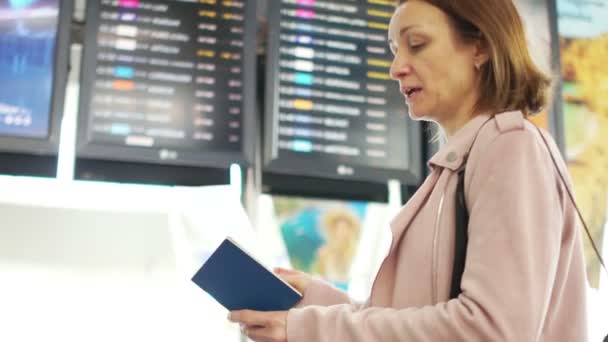 Dospělá žena sama stojí poblíž letového řádu. Podívejte se na informační tabuli odletů, přílety letadel na letiště a na letenku vybírá a porovnává s výstupním číslem — Stock video