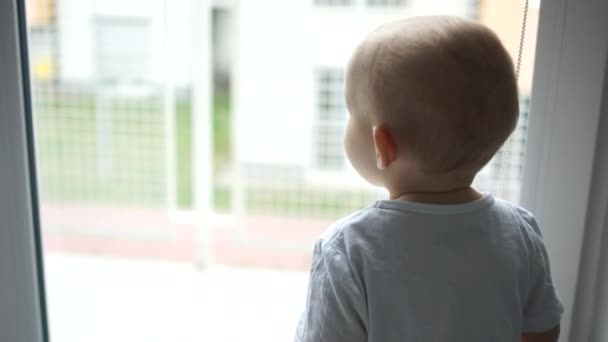 Il bambino di due anni con una maglietta bianca da solo guarda attentamente fuori dalla finestra e dice qualcosa. Figlio unico, orfanotrofio, adozione di bambini — Video Stock