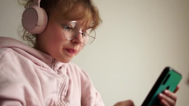 ピンクのヘッドフォンと彼女の手にスマートフォンで美しいティーンエイジャーの女の子の肖像画。十代の若者は音楽を聞く — ストック動画