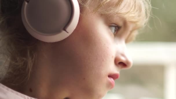 Close portrait of a girl in pink headphones by the window. Schoolgirl listens to music and is sad — 비디오