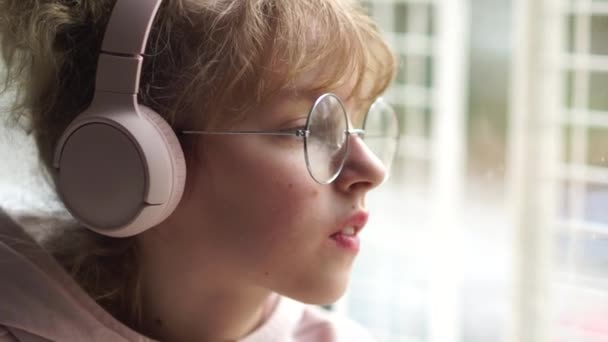 A estudante ouve música e está triste. Fechar retrato de uma menina com óculos em fones de ouvido rosa pela janela — Vídeo de Stock