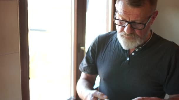 Viejo lavándose los dientes delante del espejo. Un hombre de barba gris con gafas aprieta la pasta de un tubo en un cepillo. Higiene matinal — Vídeos de Stock