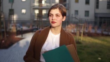 Kahverengi ceketli kısa saçlı kız elinde kağıtlarla bir dosya tutuyor. Gün batımında bir ofis binasının avlusunda genç bir iş kadını.