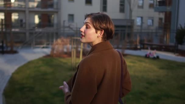 Joven empresaria en el patio de un edificio de oficinas al atardecer. Una chica de pelo corto con un abrigo marrón sostiene una carpeta con papeles en las manos — Vídeos de Stock