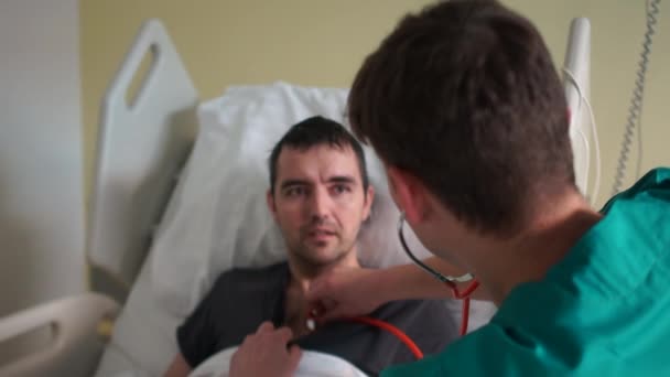 Le docteur examine un patient au lit. Écoute le cœur et les poumons avec un stotoscope. Jeune homme dans un hôpital. Coronovirus, épidémie d'Orvi — Video