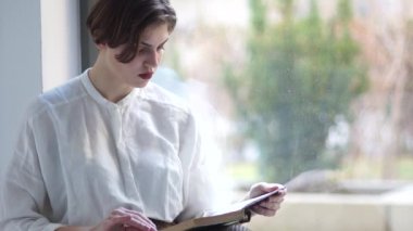 Beyaz bluzlu üzgün kız elinde bir kitapla pencerenin üzerinde oturuyor. Tatildeki kız öğrenci, mutlu kadın.